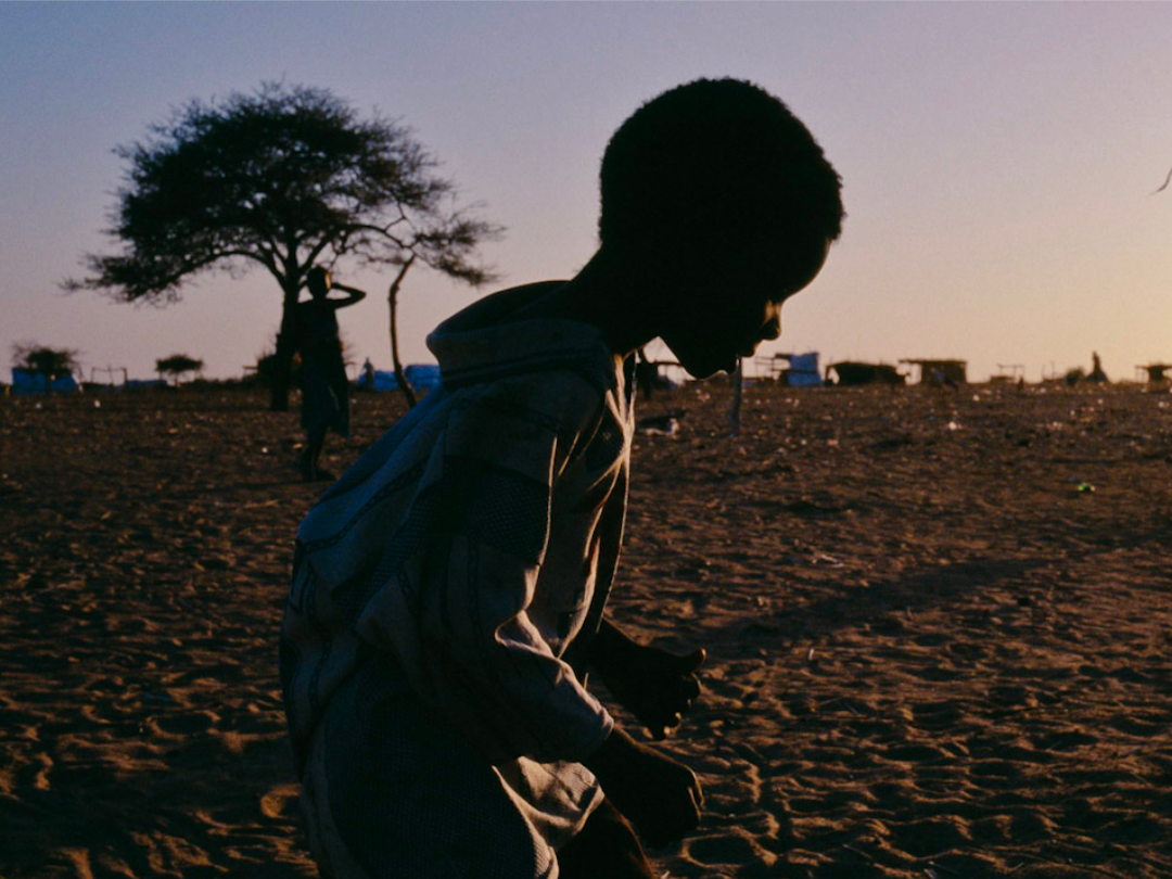 Silhouette of a child crosses in front of the camera with a silhouette of a woman carrying a bundle on her head in the background (Darfur, Sudan)