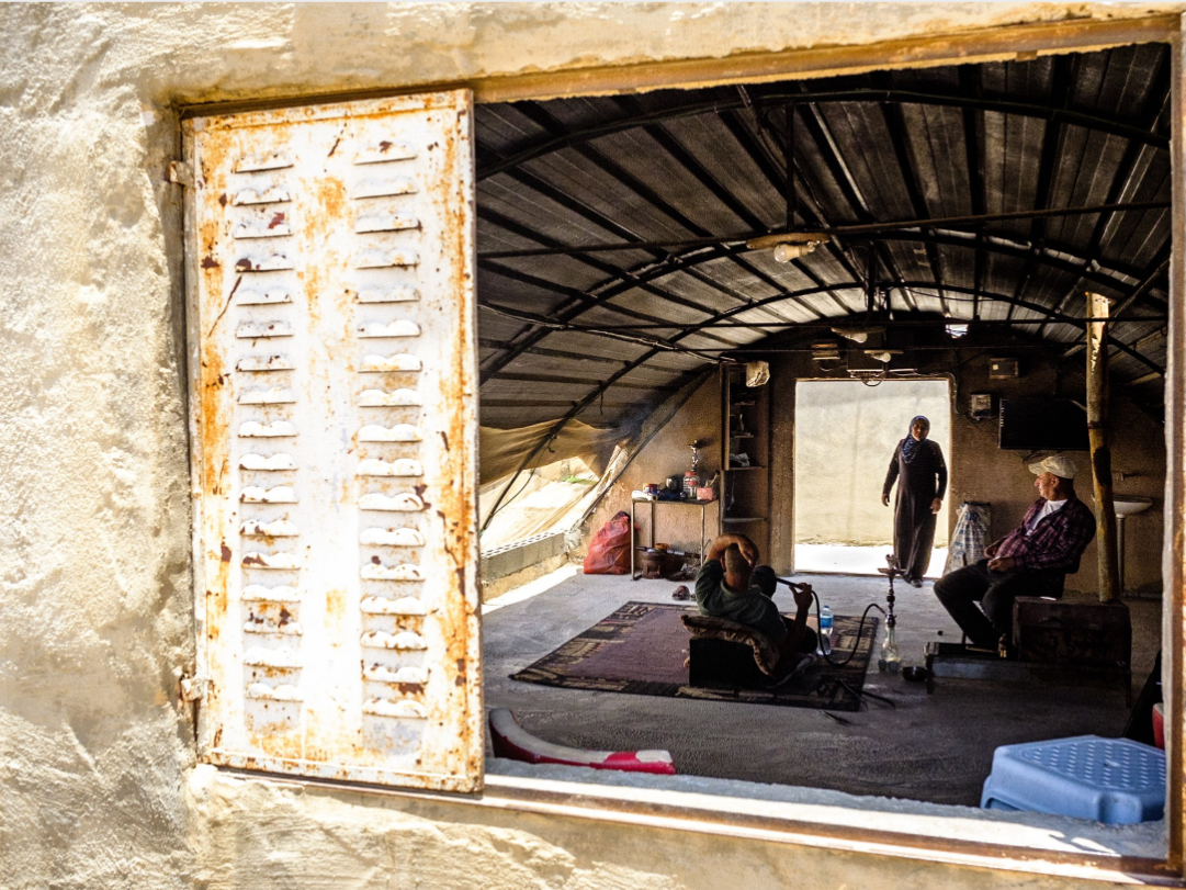 A window looks in to a curved roofed structure, with people sitting and a woman standing in the far doorway