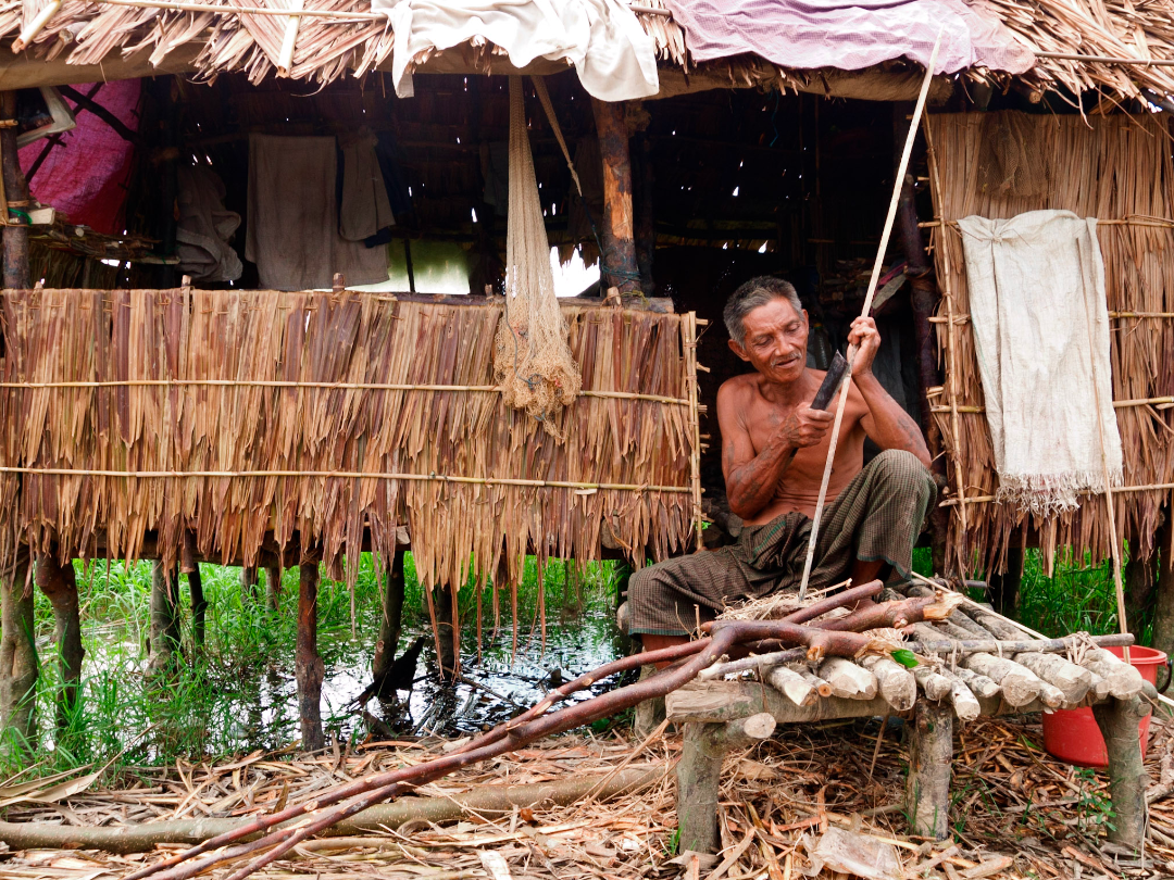 An elderly Myanmar man whittles sticks in front of a reed house (Irrawaddy Delta)